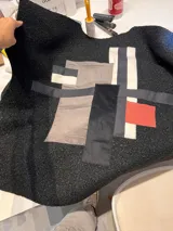 Campera de paño negro con diseño de collage geométrico aplicado en el frente, compuesto por parches de tela en tonos gris, blanco, negro y un detalle en rojo. Presenta costuras a la vista y una textura rugosa característica del paño.