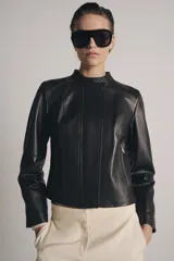 Chaqueta de cuero de corte entallado con cuello subido, cierre frontal con cremallera y botones ocultos bajo solapa. Presenta bolsillos delanteros con cierre y mangas largas terminadas en cremalleras metálicas.