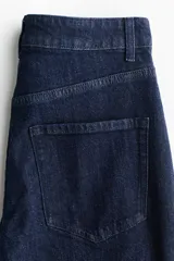 Pantalón de jean de denim azul oscuro, de tiro alto y piernas amplias con corte holgado. Presenta solapas de bolsillo decorativas con botón en el frente y costuras verticales visibles a lo largo de las piernas.