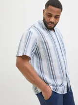 Camisa de manga corta con cuello resort y ajuste relajado. Presenta un diseño de rayas verticales en tonos azules y blancos, con cierre frontal de botones. Confeccionada en una mezcla de algodón y lino, ofrece una textura ligera y fresca.