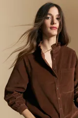 Camisa de pana color marrón, con cuello alto decorado con volados, cierre frontal con botones y bolsillo aplicado en el pecho. Presenta mangas largas con puños ajustados.