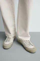 Zapatilla deportiva de estilo retro, color blanco con detalles en gris y negro. Presenta una suela de color beige en contraste y cordones blancos.