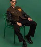 Campera bomber de estilo college con cuerpo a cuadros en tonos marrones y mangas largas de cuero sintético negro. Presenta cuello, puños y cintura acanalados a rayas, cierre abotonado, bolsillos laterales y aplique bordado de letra "B" en el pecho.
