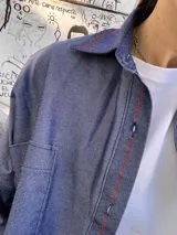 Sobre camisa de gabardina símil denim, color azul, con costuras visibles en color rojo y etiqueta de cuero en la espalda. Presenta un corte holgado y mangas largas.