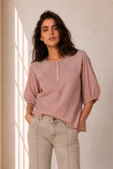 Blusa a rayas verticales en tonos rosados y blancos, con cuello redondo y cierre frontal parcial con botones. Presenta delicados detalles bordados en los hombros y mangas tres cuartos abullonadas con puños elásticos.