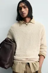 Jersey de punto color beige claro, con textura irregular y cuello redondo. Presenta un calce relajado y manga larga con puños y ruedo en rib.