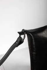 Cartera bandolera de cuero negro con flecos colgantes en el lateral, cierre superior y correa ajustable con argollas metálicas.
