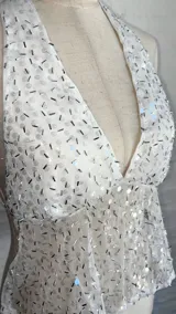Top halter blanco con escote pronunciado en V, confeccionado en tejido transparente con lentejuelas o brillos plateados. La parte superior del busto es opaca y el resto del cuerpo es traslúcido.