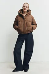 Campera puffer acolchada de color negro, con cuello envolvente, capucha integrada, manga larga, bolsillos laterales ocultos y bajo con elástico. Cierre frontal con botones a presión ocultos por solapa.