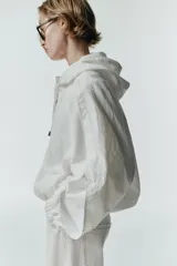 Chaqueta blanca de cuello redondo con capucha ajustable mediante cordones elásticos. Presenta manga larga con puños elásticos, bolsillos delanteros y bajo asimétrico ajustable con cordones y stoppers. El cierre frontal combina cremallera y botones ocultos bajo una solapa.