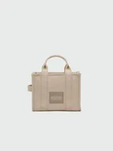 Bolso tipo tote de lona color beige de la marca Marc Jacobs. Presenta el texto 'THE TOTE BAG' estampado en el frente en color blanco, dos asas de mano superiores y una correa de hombro ajustable y removible en color negro.