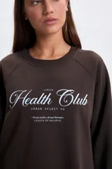 Buzo deportivo de corte oversize en color marrón, con cuello redondo y mangas largas tipo raglán. Presenta una estampa frontal con la inscripción 'Health Club' en tipografía cursiva y detalles de texto en blanco.