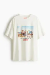 Remera oversized de punto de algodón color blanco, con cuello redondo y mangas cortas de hombros caídos. Presenta un diseño estampado en el pecho con temática fotográfica retro y la inscripción "Fiesta Hollywood Party, 1974 Pacha".