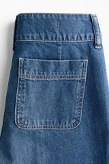 Pantalón de jean estilo wide leg con tiro alto, confeccionado en denim de algodón rígido. Presenta bolsillos delanteros con detalle de ribete trenzado, pliegues frontales y traseros, y cierre con doble botón.