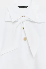 Camisa blanca de cuello solapa y manga larga, con detalle de lazada en el mismo tejido en la parte frontal y cierre con botones dorados.