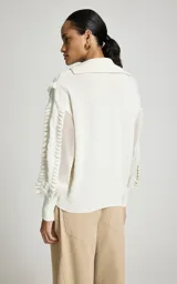 Sweater calado de punto color blanco, de corte recto con amplio cuello tipo polo y escote en V. Presenta mangas largas con volumen, puños ajustados y detalles de volados texturizados en la parte delantera y en las mangas.