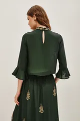 Camisa de viscosa color verde oscuro, con cuello alto y mangas tres cuartos terminadas en volados. Presenta un elaborado bordado artesanal en el pecho con pedrería dorada y detalles decorativos en la cintura.