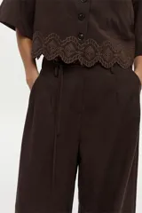Camisa de manga corta color marrón, con cuello solapa, cierre frontal de botones, bolsillo en el pecho y detalle de bordado decorativo en el ruedo con terminación ondulada.