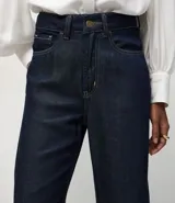 Pantalón de corte recto y cintura alta, confeccionado en denim crudo (raw denim) de color azul oscuro. Presenta cierre frontal con botón y cremallera, trabillas para cinturón y bolsillos funcionales en frente y espalda.