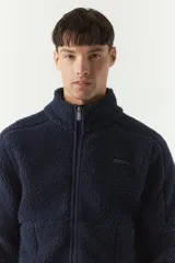 Campera de sherpa color verde militar, con cierre frontal completo, cuello alto y bolsillos laterales con cierre. Presenta un parche pequeño con logo en el pecho.