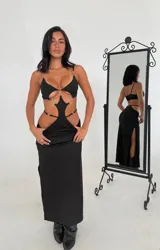 Vestido largo negro con aberturas cut-out en el abdomen y la espalda, decorado con aros metálicos plateados. Tiene tirantes finos y una falda larga con abertura lateral.