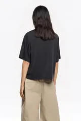 Camiseta de manga corta y escote redondo, color beige claro, con corte boxy. Presenta una hilera de tachuelas metálicas negras decorativas en la parte superior del pecho y en las costuras de los hombros.