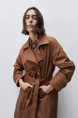Trench clásico color marrón, de silueta relajada, confeccionado en tejido liviano, con cuello de solapa, doble abotonadura frontal y mangas largas con presillas regulables en los puños.