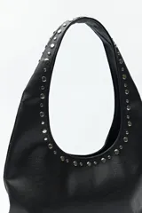 Cartera tipo hobo de símil cuero texturizado color marrón oscuro, con apliques de tachas metálicas plateadas que recorren el frente y el contorno. Cuenta con un bolsillo frontal y un asa de hombro.