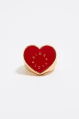 Anillo dorado con forma de corazón rojo y letras doradas.