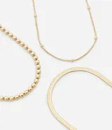 Conjunto de tres collares de metal con baño dorado claro. Incluye un collar de cadena plana tipo cinta italiana, un collar de cuentas esféricas pequeñas y un collar de cuentas tipo media esfera. Fabricados en una mezcla de latón y hierro reciclado.