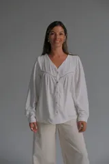 Blusa blanca de corte holgado con cuello en V, cierre frontal con botones y detalles de alforzas y encaje en el pecho. Presenta mangas largas con puños rectos.