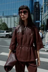 Chaqueta efecto piel color marrón, con cuello solapa, mangas por debajo del codo, cintura elástica, falsos bolsillos delanteros y cierre frontal con cremallera.