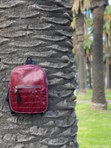 Mochila color bordó con textura tipo reptil en la parte superior y bolsillo frontal con diseño capitoneado en material charolado. Cuenta con cierre metálico y tiradores con flecos decorativos.