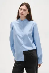 Camisa celeste de manga larga con cuello alto y volados, detalles de frunces en hombros y puños, y cierre frontal con botones.