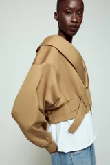 Chaqueta corta de Zara confeccionada en mezcla de algodón, con cuello solapa, manga larga con hombro caído y puños elásticos. Presenta un diseño cruzado con cierre de hebilla metálica lateral y forro interior a tono.