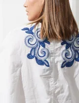 Blusa blanca con bordado floral azul, escote en V, manga japonesa y cierre frontal con botones.