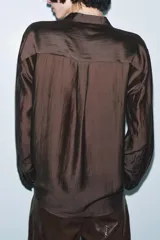 Camisa fluida color marrón oscuro, con escote en pico y cuello tipo solapa. Manga larga con puños anchos y bajo asimétrico. Cierre frontal con botones ocultos.