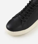 Championes casuales para hombre confeccionados en cuero negro, con diseño minimalista de caña baja, detalles en color marrón en el talón, cordones elásticos y suela alta de goma en tono off-white.