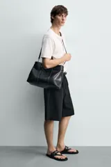 Bolso de mano estructurado en color negro, con diseño minimalista y detalle de cinturón decorativo en la parte superior. Cuenta con doble asa de mano y cierre magnético.