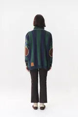 Buzo de punto con diseño de rayas verticales en tonos verde y azul marino. Presenta cuello alto con cierre frontal, hombros caídos y coderas en contraste de color marrón con costuras decorativas visibles.