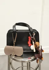 Funda para lentes color beige con textura símil cuero, cierre con broche a presión y correa con mosquetón para colgar de la cartera.