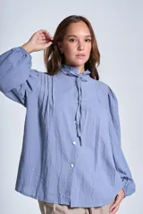 Camisa de color azul con cuello alto decorado con volados y lazo de ajuste. Presenta tablas decorativas en los hombros, cierre frontal con botones de nácar y mangas largas con puños elásticos que generan un efecto abullonado.