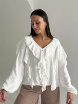 Blusa blanca de corte holgado con escote en V, decorada con volados frontales y en las mangas. Presenta mangas largas con puños elásticos y cierre frontal con botones.