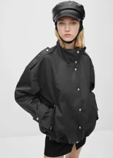 Anorak de diseño oversize en color negro, con cuello alto, cierre frontal de botones y cremallera oculta bajo tapeta. Presenta mangas largas con puños de tira abotonada, bolsillos laterales con cremallera y detalles de charreteras en los hombros.