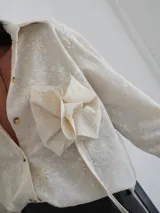 Camisa color crema con bordado de flores y roseta decorativa en el frente. Tiene botones de cuerno, corte holgado y espalda más larga.