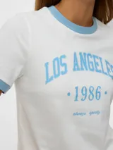Remera blanca de manga corta con cuello redondo y ribetes celestes en cuello y mangas. Estampa frontal con el texto "LOS ANGELES •1986•" en celeste.