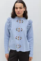 Sweater de lana color celeste, con cuello redondo, mangas largas y volados decorativos en los hombros. Presenta bordados florales artesanales en el frente y detalles de tejido calado vertical.
