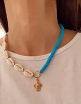 Collar de diseño asimétrico compuesto por una mitad de caracolas naturales y una mitad de cuentas de silicona color turquesa, con un dije central de tortuga en tono dorado.