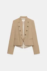 Blazer entallado de sarga color beige con cuello subido y escote en pico. Presenta manga larga con hombreras, detalle de botones metálicos decorativos en el frente y cierre frontal con gancho metálico. Incluye falsos bolsillos de vivo y abertura en la espalda.