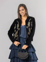 Chaqueta de terciopelo negro con bordados florales y ornamentales en tonos dorados, rosados y azules. Presenta un corte recto, cuello redondo y cierre frontal abierto con detalles de costuras decorativas en los bordes.